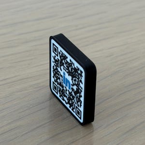 May include: A small, rectangular, black-edged plaque featuring a white face with a black QR code pattern and a blue LinkedIn logo in the center. The object is positioned at an angle, displaying its thickness and the printed graphic design clearly.