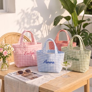 Puede incluir: Cuatro bolsos de tela pequeños con estampado de cuadros vichy en rosa, azul, verde y rojo. El bolso rosa está bordado con el nombre Evelyn, el azul con Aurora y el verde con Victoria. Cada bolso cuenta con dos asas y una abertura superior.