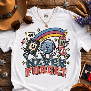 Könnte beinhalten: Ein weißes T-Shirt mit einer bunten Grafik aus einer Zeichentrick-Kassette, einem CD-Player und einem Klapphandy unter einem Regenbogen. Der Text "NEVER FORGET" steht in großen, abgenutzten blauen und roten Buchstaben unter den Figuren.
