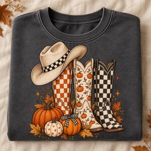 Puede incluir: Una sudadera de cuello redondo gris carbón con un estampado gráfico de un sombrero de vaquero beige sobre tres botas vaqueras. Las botas muestran patrones de cuadros naranjas, enredaderas de calabaza y cuadros negros, rodeadas de calabazas.