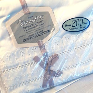 May include: A white Wamsutta Supercale Plus double flat sheet in plastic packaging. The sheet features a decorative eyelet lace border. The label indicates 200 thread count, 50% Fortrel polyester, 50% combed cotton, and a size of 81 x 96 inches.