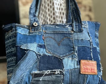 May include: A tote bag constructed from various shades of blue denim fabric patches. The bag features a central back pocket with a red Levi's tab, a tan leather brand patch, and two denim handles. The design incorporates visible seams and frayed edges.