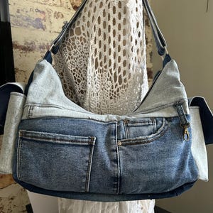Può includere: Una borsa a spalla in stile hobo realizzata in tessuto denim blu riciclato. La borsa presenta un design bicolore con pannelli in denim blu chiaro e scuro, molteplici tasche esterne, una chiusura superiore con zip e un moschettone dorato.