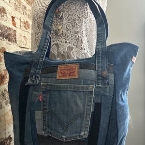 Op de afbeelding: Een draagtas gemaakt van hergebruikte blauwe spijkerstof met een centrale buitenzak, twee schouderbanden en een bruin leren merklabel van Levi Strauss & Co. Het ontwerp bevat verschillende tinten blauwe denim in diverse panelen.
