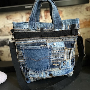 Könnte beinhalten: Eine Patchwork-Tragetasche aus verschiedenen Blautönen von Jeansstoff. Die Tasche verfügt über eine markante Jeans-Tasche, mehrere Metallnieten und ein horizontales schwarzes Stoffband. Sie umfasst zwei Tragegriffe und einen abnehmbaren schwarzen Schultergurt.