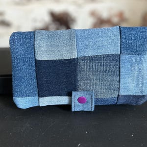 Peut inclure: Un portefeuille rectangulaire composé d'un assemblage de carrés de tissu en jean dans diverses nuances de bleu, allant du bleu clair au bleu indigo foncé. Le portefeuille présente une petite patte de fermeture en jean avec un bouton-pression violet.