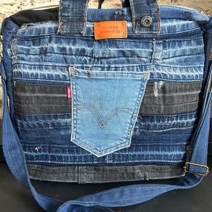 Op de afbeelding: Een patchwork spijkerstof tas gemaakt van verschillende tinten blauwe en zwarte denim stof. De tas heeft een lichtblauw middenvak met een rood Levi's label, een oranje leren label met de tekst Ax Yo Nel Denim & Co en een metalen knoop.