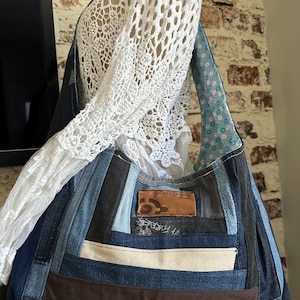 May include: A hobo-style shoulder bag constructed from patchwork denim strips in various shades of blue, brown, and cream. The bag features a rectangular brown leather patch with a circular logo and the text "JEANS" and a light blue floral lining.