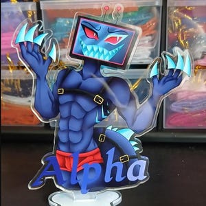 Peut inclure: Un présentoir en acrylique transparent représentant un personnage bleu musclé avec un écran de télévision en guise de tête, des yeux rouges et des dents pointues. Le personnage porte un slip rouge et des sangles noires, avec le mot Alpha en bleu.