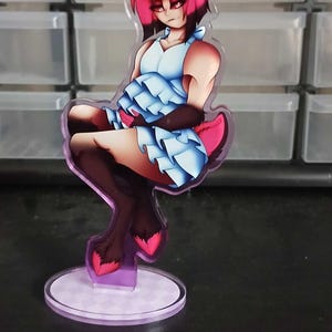 Peut inclure: Une figurine en acrylique représentant un personnage stylisé aux cheveux rose vif, aux oreilles sombres et à la fourrure brune, portant une robe à volants bleu clair. Le personnage est assis sur un socle circulaire à motif à carreaux violet.