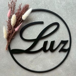 Op de afbeelding: Een zwart cirkelvormig wandbord met de naam Luz in een cursief lettertype. Een klein boeket gedroogde bloemen in roze, crème en bruine tinten is bevestigd aan de linkerkant van de zwarte cirkelvormige lijst, wat zorgt voor een natuurlijk accent.