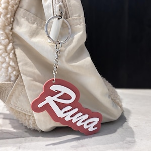 Può includere: Un portachiavi personalizzato con il nome Runa in caratteri corsivi bianchi su uno sfondo color rosa polvere. Il portachiavi è fissato a un anello e una catenella in metallo argentato, agganciati alla cerniera di uno zaino in tessuto beige testurizzato.