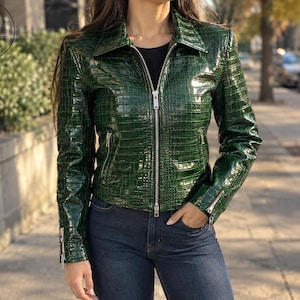 May include: A glossy dark green jacket with a textured crocodile-embossed pattern, featuring a silver-toned front zipper closure, a pointed collar, and zippered pockets on the sides and cuffs. The jacket is worn over a plain black top and blue jeans.