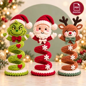 May include: Three crochet figures on spiral bases: a green character with a red Santa hat and heart accents, a Santa Claus with a red hat, and a brown reindeer with antlers and a green bow. Each figure stands on a round base.