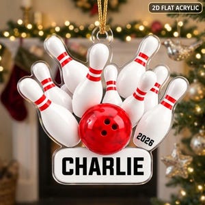 May include: A 2D flat acrylic hanging ornament featuring a red bowling ball and several white bowling pins with red stripes. The ornament includes the text 2026 and the name CHARLIE printed in black on a white base, suspended by a gold cord.
