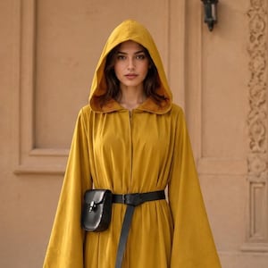 Pode incluir: Uma túnica longa até ao chão de cor amarelo mostarda com capuz, mangas largas e acampanadas e fecho de correr frontal. Um cinto de couro preto com uma pequena bolsa retangular presa está ajustado à cintura. Peça de cor sólida com capuz.