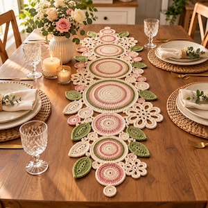 Puede incluir: Un camino de mesa decorativo de ganchillo largo que presenta una serie de círculos concéntricos en tonos crema, rosa empolvado y verde salvia, acentuado con motivos de hojas y flores de ganchillo, sobre una mesa de comedor de madera.