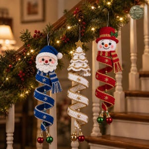 May include: Three crocheted Christmas ornaments hanging from a garland. One features a blue Santa, one a white Christmas tree with gold accents, and one a snowman wearing a red hat and scarf. Each has a spiral tail ending in two small round bells.