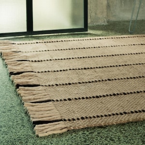 May include: A rectangular woven rug featuring a textured beige and tan surface with horizontal dark brown stripes and thick, knotted fringe detailing along the edges. The rug is positioned on a green floor near a metal wire chair.