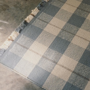 May include: A rectangular area rug featuring a large-scale plaid pattern in shades of dusty blue and cream. The rug has a woven texture and is finished with short, decorative fringe along the edge, alternating between blue and cream colored threads.
