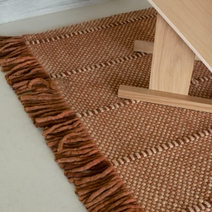 May include: A rectangular rug in a warm terracotta hue featuring a textured, woven pattern with thick, knotted fringe along the edge. A light-toned wooden chair with a brown leather headrest is positioned partially over the rug.