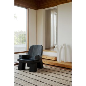 May include: A black armchair with a low back, wide armrests, and four block-shaped legs. Beside the chair are three tall, white, slender, abstract sculptures with rounded tops, positioned on a wooden platform near a window.