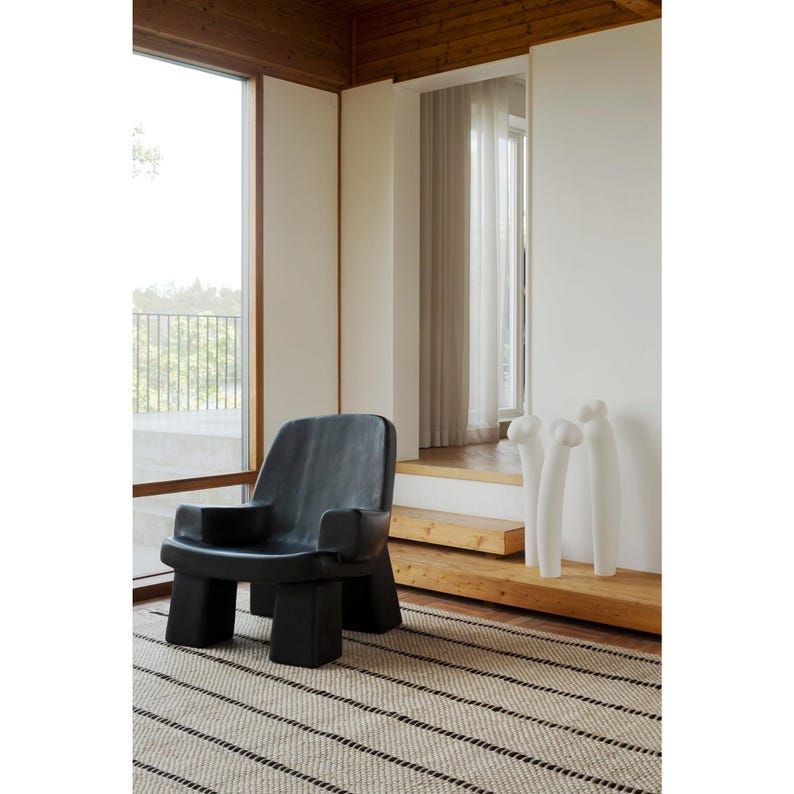 May include: A black armchair with a low back, wide armrests, and four block-shaped legs. Beside the chair are three tall, white, slender, abstract sculptures with rounded tops, positioned on a wooden platform near a window.