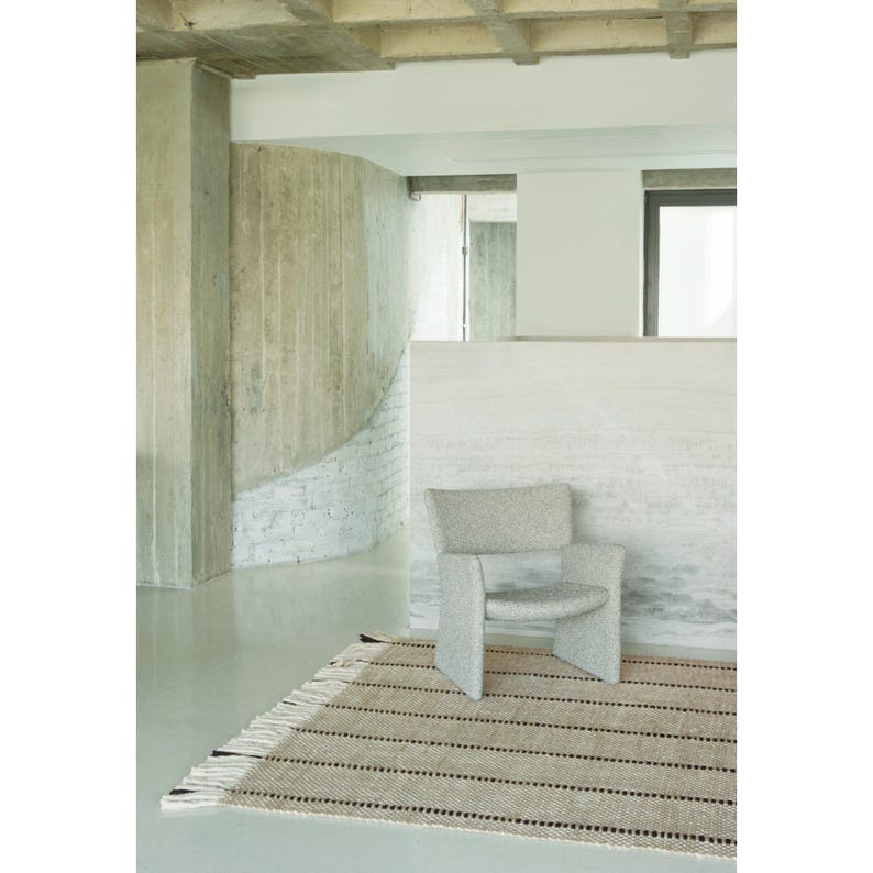 May include: A light gray upholstered armchair with a curved backrest and wide armrests, positioned on a rectangular woven rug featuring a pattern of horizontal tan and black stripes with white fringe detailing on the short ends.
