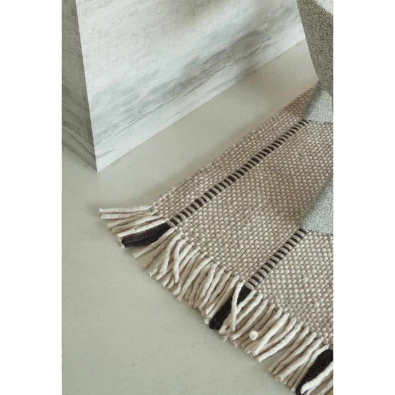 May include: A rectangular woven rug featuring a textured beige and cream surface with vertical dark brown stripes. The rug is finished with long, thick fringe tassels along the edge, alternating between cream and dark brown yarn strands.