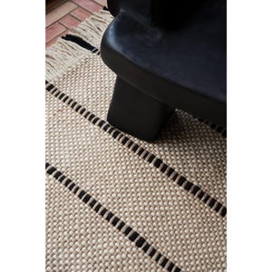 May include: A woven rug featuring a textured beige and cream pattern with horizontal black stripes and a fringed edge, positioned beneath a smooth, matte black furniture piece with a rounded, solid base structure.
