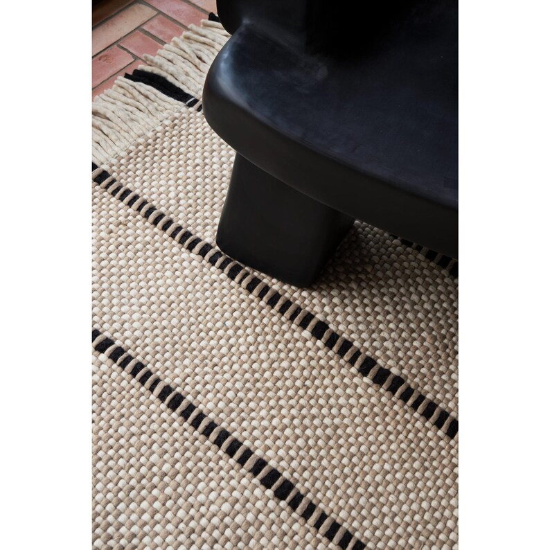 May include: A woven rug featuring a textured beige and cream pattern with horizontal black stripes and a fringed edge, positioned beneath a smooth, matte black furniture piece with a rounded, solid base structure.
