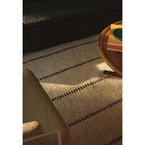 May include: A rectangular area rug featuring a textured, cream-colored woven surface with evenly spaced, horizontal dark brown stripes. The rug is positioned on the floor, partially covered by a wooden table and a chair.