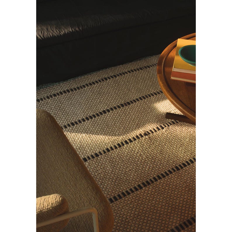 May include: A rectangular area rug featuring a textured, cream-colored woven surface with evenly spaced, horizontal dark brown stripes. The rug is positioned on the floor, partially covered by a wooden table and a chair.