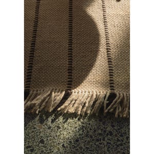 May include: A woven rug featuring a textured cream-colored surface with vertical dark brown stripes. The rug is finished with a fringe of thick, cream-colored yarn tassels along the bottom edge, creating a tactile and layered appearance.