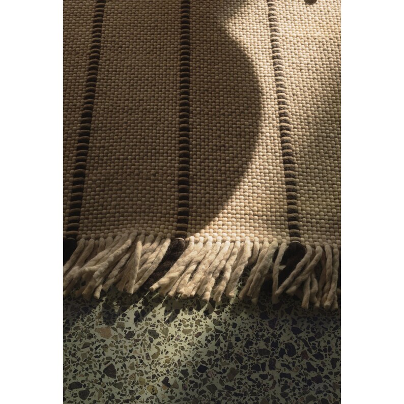 May include: A woven rug featuring a textured cream-colored surface with vertical dark brown stripes. The rug is finished with a fringe of thick, cream-colored yarn tassels along the bottom edge, creating a tactile and layered appearance.