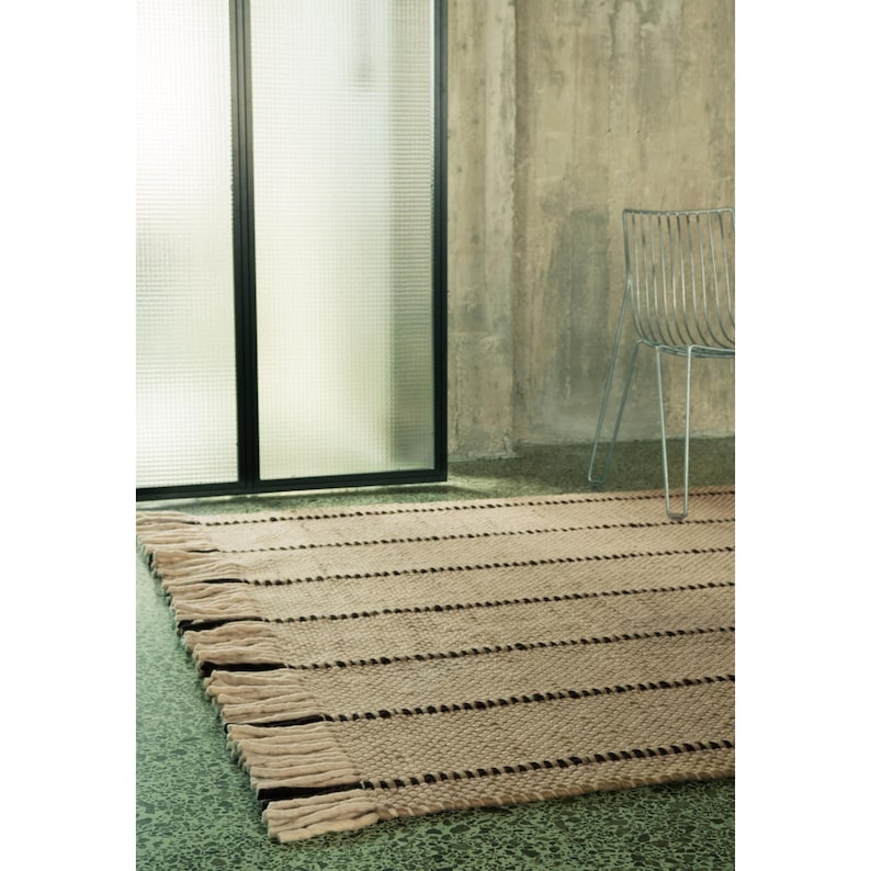 May include: A rectangular woven rug featuring a textured beige and tan surface with horizontal dark brown stripes and thick, knotted fringe detailing along the edges. The rug is positioned on a green floor near a metal wire chair.