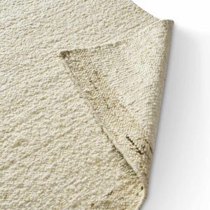 May include: A cream-colored area rug with a thick, textured, high-pile surface. The rug is folded over at one corner, revealing the underside of the fabric, which features a visible woven structure and subtle dark brown flecks throughout the weave.