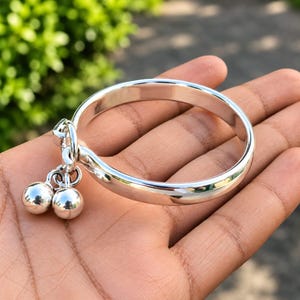 May include: A polished silver-tone bangle bracelet featuring a smooth, rounded band and two small, spherical bell charms attached to a single jump ring. The metal surface is reflective, showing highlights and a bright, metallic finish throughout.