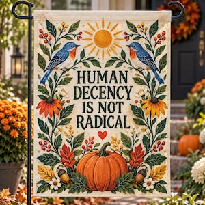 May include: A rectangular garden flag featuring the text HUMAN DECENCY IS NOT RADICAL in dark green letters. The design includes two blue birds, a yellow sun, orange flowers, green leaves, red berries, a large orange pumpkin, and a small red heart.