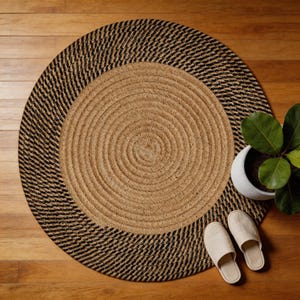 May include: A round braided rug featuring a natural tan center and a wide black and tan outer border. The rug is constructed from coiled fibers, creating a textured, circular pattern throughout the surface of the floor covering.