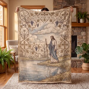 May include: A rectangular throw blanket featuring a printed illustration of a figure in robes holding a staff by a river. The design includes grapevines, crosses, and the text "HE LEADETH ME BESIDE THE STILL WATERS" in a banner at the top.