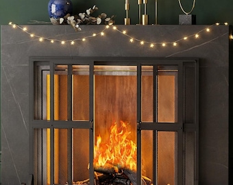 May include: A black metal fireplace screen with a three-panel design and fine mesh detailing. The screen features a sturdy frame with curved feet, positioned in front of a stone fireplace hearth containing burning logs and bright orange flames.