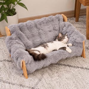 May include: A rectangular pet bed featuring a plush, grey faux fur cushion supported by a light-colored wooden frame. A white and grey tabby cat is lying down on the soft, textured surface of the bed, which is elevated by four wooden legs.