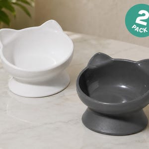 May include: A 2-pack of ceramic pet bowls featuring a cat-ear silhouette. One bowl is solid white and the other is solid dark gray. Each bowl has a wide, circular base and a smooth, glossy finish, designed with a raised rim shaped like cat ears.