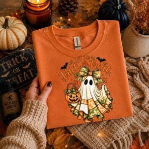 Può includere: Una felpa girocollo arancione con la grafica di un fantasma che indossa un fiocco mimetico e tiene in mano un secchiello a forma di zucca. Il testo recita Off To Trick or Treat, accompagnato da illustrazioni di pipistrelli e foglie autunnali.