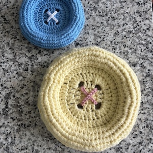 May include: Two circular crochet items shaped like large buttons. One is light blue with white yarn stitched in an X pattern in the center. The other is pale yellow with pink yarn stitched in an X pattern in the center. Both have a raised rim.