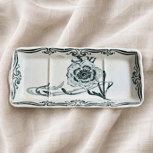 May include: A rectangular white ceramic tray featuring a dark green floral illustration in the center and decorative scrollwork along the border. The tray has two vertical indentations on the surface and a slightly wavy, raised edge.