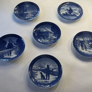 May include: Five circular porcelain plates featuring blue and white winter scenes. Each plate displays a unique landscape illustration, including a water mill, a train, a hunter with a dog, a family with a tree, and snowy mountain vistas.