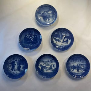 May include: A collection of five circular blue and white porcelain decorative plates, each featuring a distinct winter scene and a specific year printed along the bottom rim, including 1980, 1981, 1985, 1986, and 1987, with detailed illustrations.