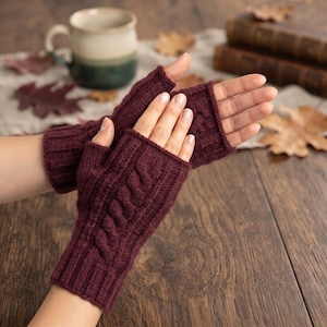 May include: A pair of fingerless gloves in a deep burgundy color, featuring a vertical cable knit pattern on the back of the hand and a ribbed cuff at the wrist. The gloves are crafted from a soft knit material and leave the fingers exposed.